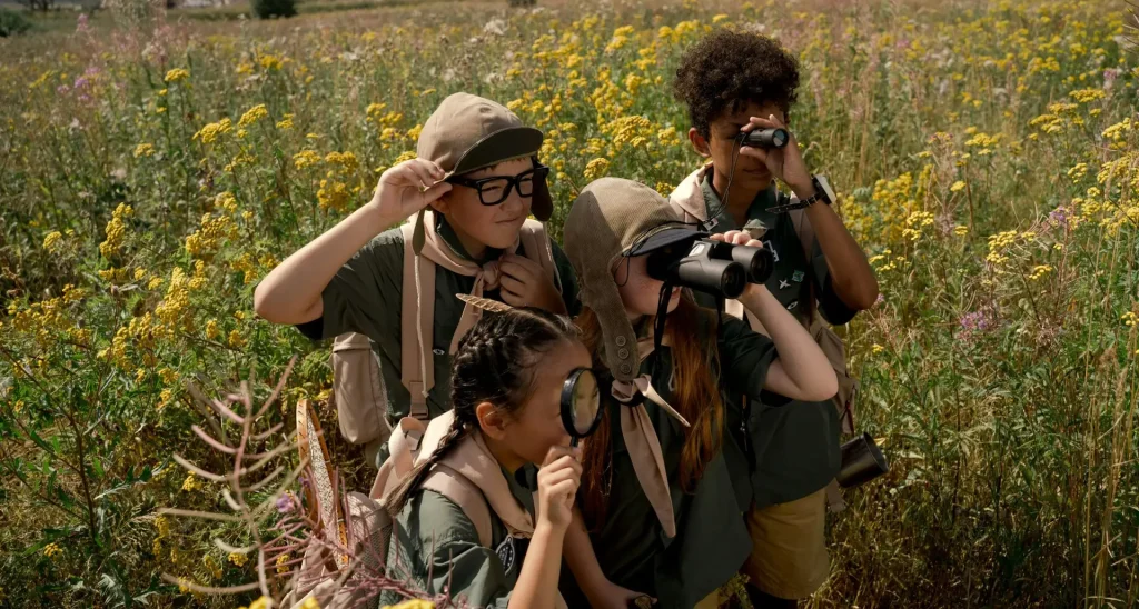 Group of child scouts exploring a meadow with binoculars and magnifying glass on a sunny day.