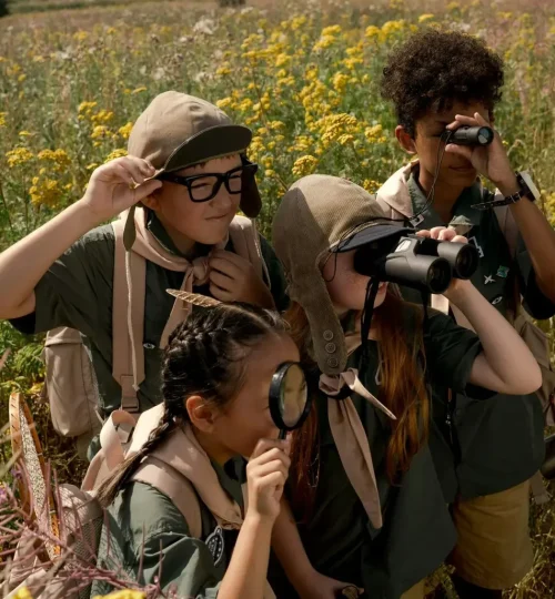 Group of child scouts exploring a meadow with binoculars and magnifying glass on a sunny day.