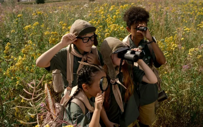 Group of child scouts exploring a meadow with binoculars and magnifying glass on a sunny day.